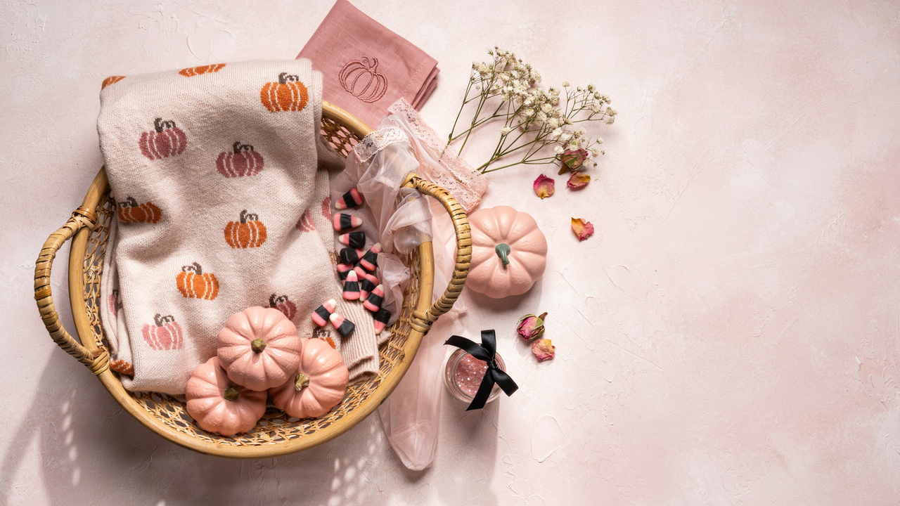 A top-down photo of a wicker basket filled with a cream-colored pumpkin-patterned blanket, pink ceramic pumpkins, and candy corn, set against a textured light pink background with baby's breath and dried rose petals.