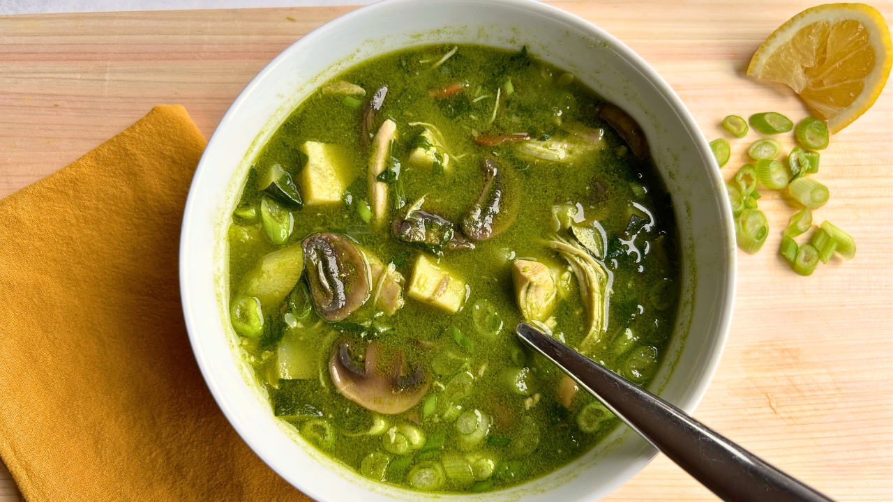 White ceramic bowl of Lemongrass Ginger Breakfast Soup with chicken, mushrooms, zucchini, spinach, green onions, and a lemon wedge, on a wood cutting board with a spoon and a mustard yellow napkin.