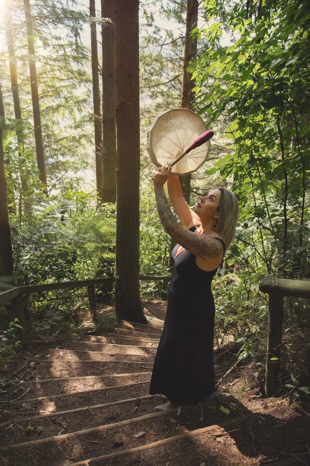 Victoria Bond raising a frame drum on a sunlit forest path