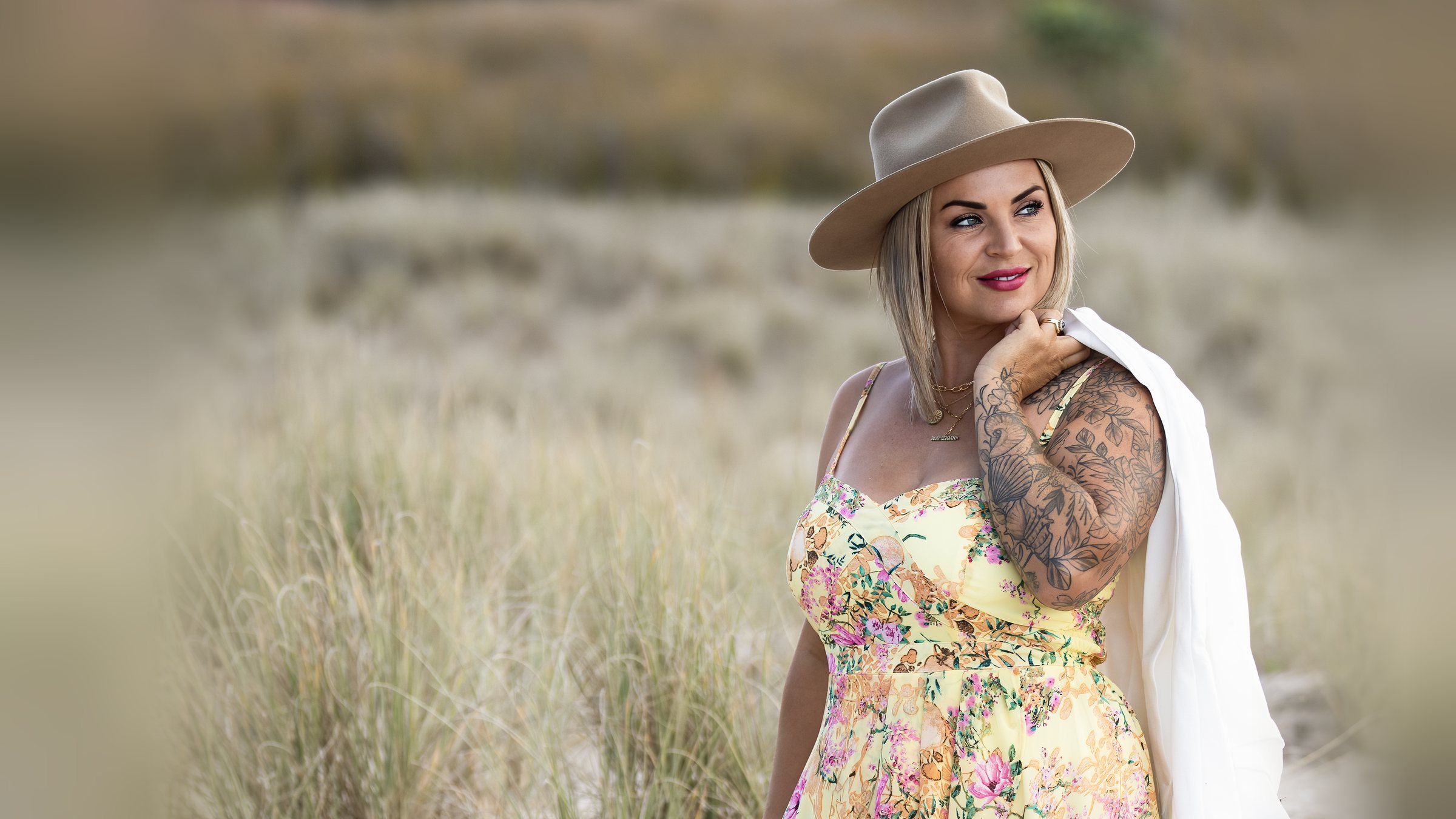 Victoria Bond smiling in a floral dress and wide brim hat, a white jacket over her shoulder, in the dune grass