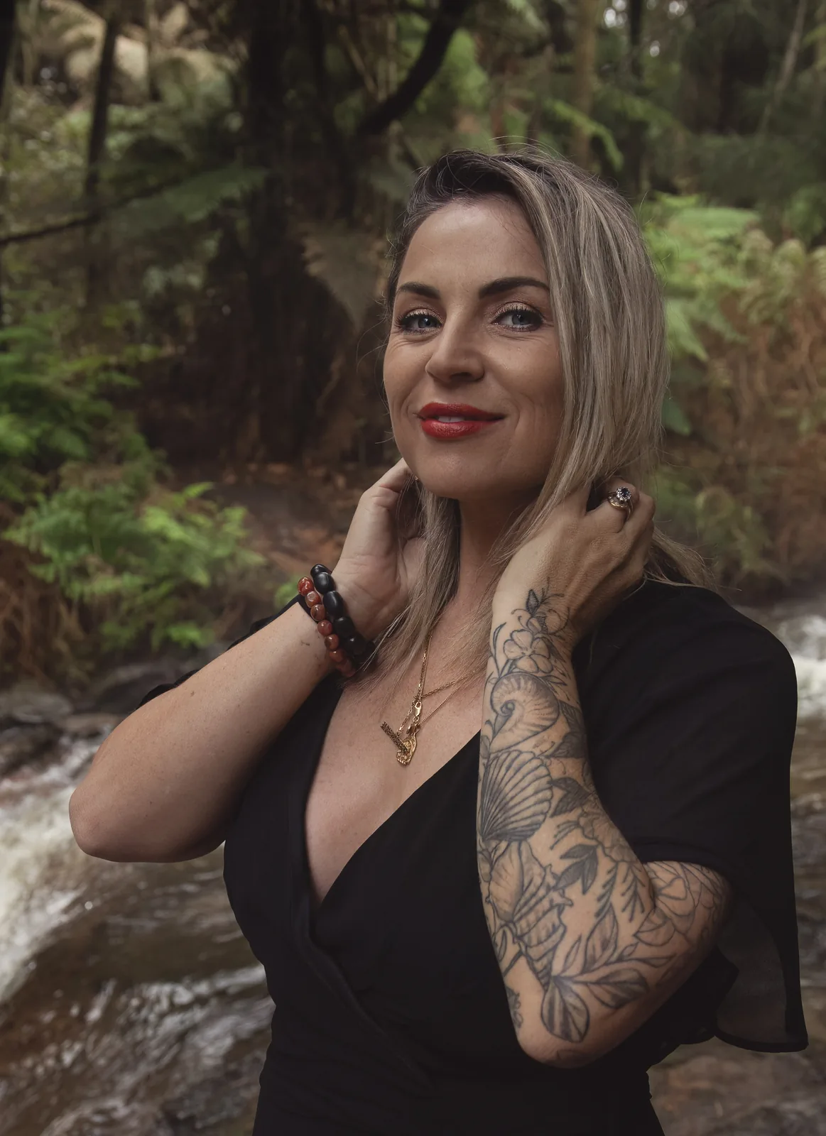 Victoria Bond smiling, hands at her neck, among ferns beside a forest stream