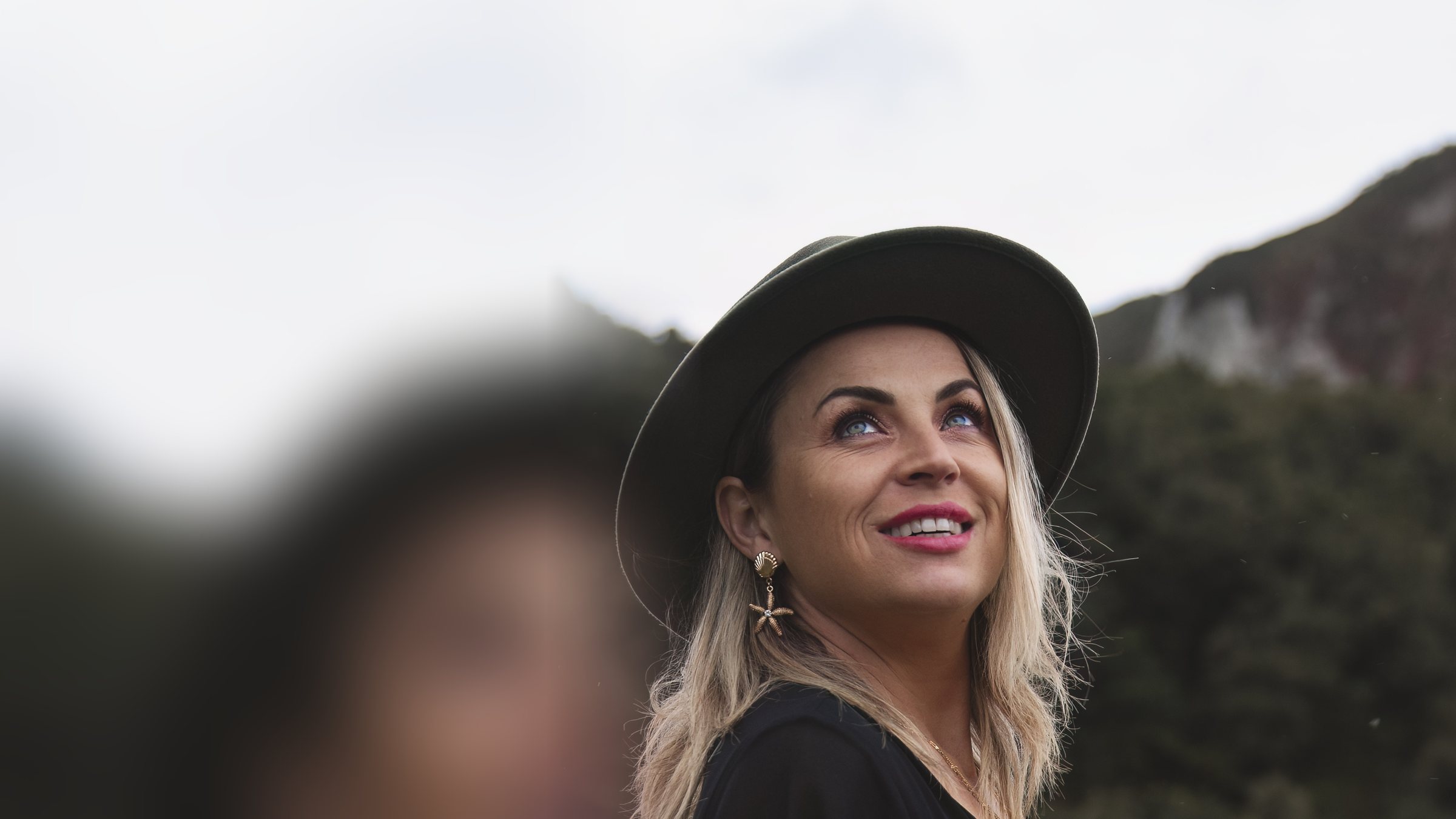 Victoria Bond in a dark hat, looking up with an open smile, bush-covered cliffs behind her