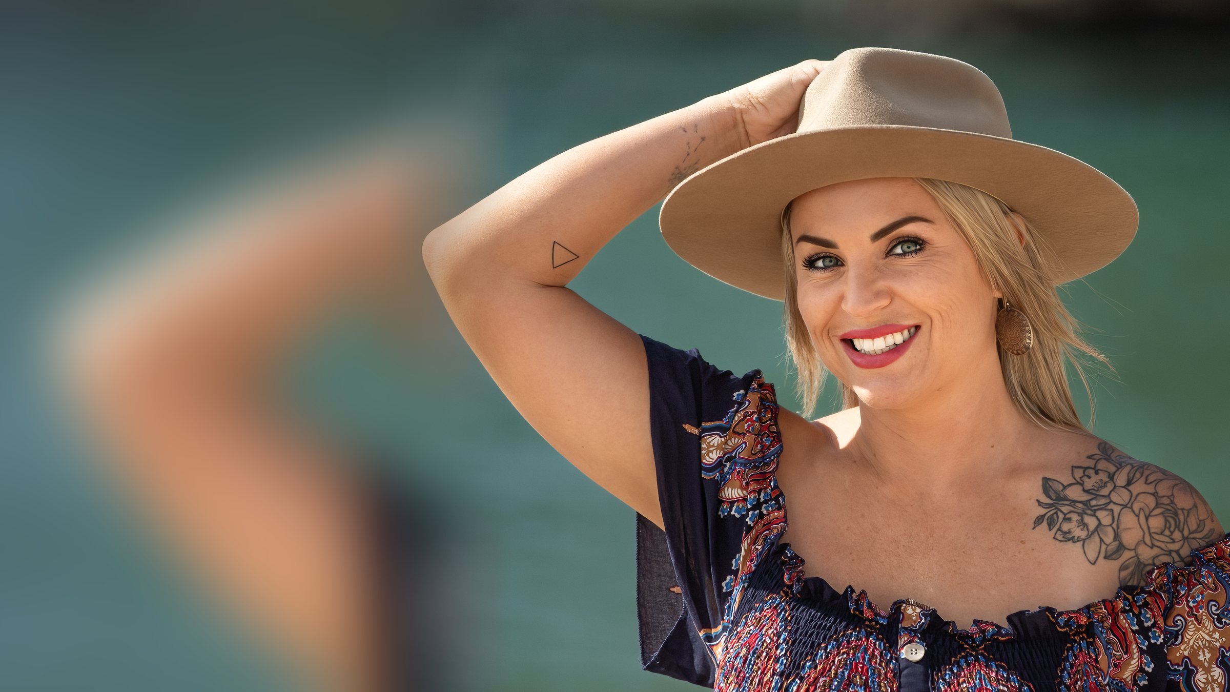 Victoria smiling warmly, one hand on her hat, with soft teal water behind her
