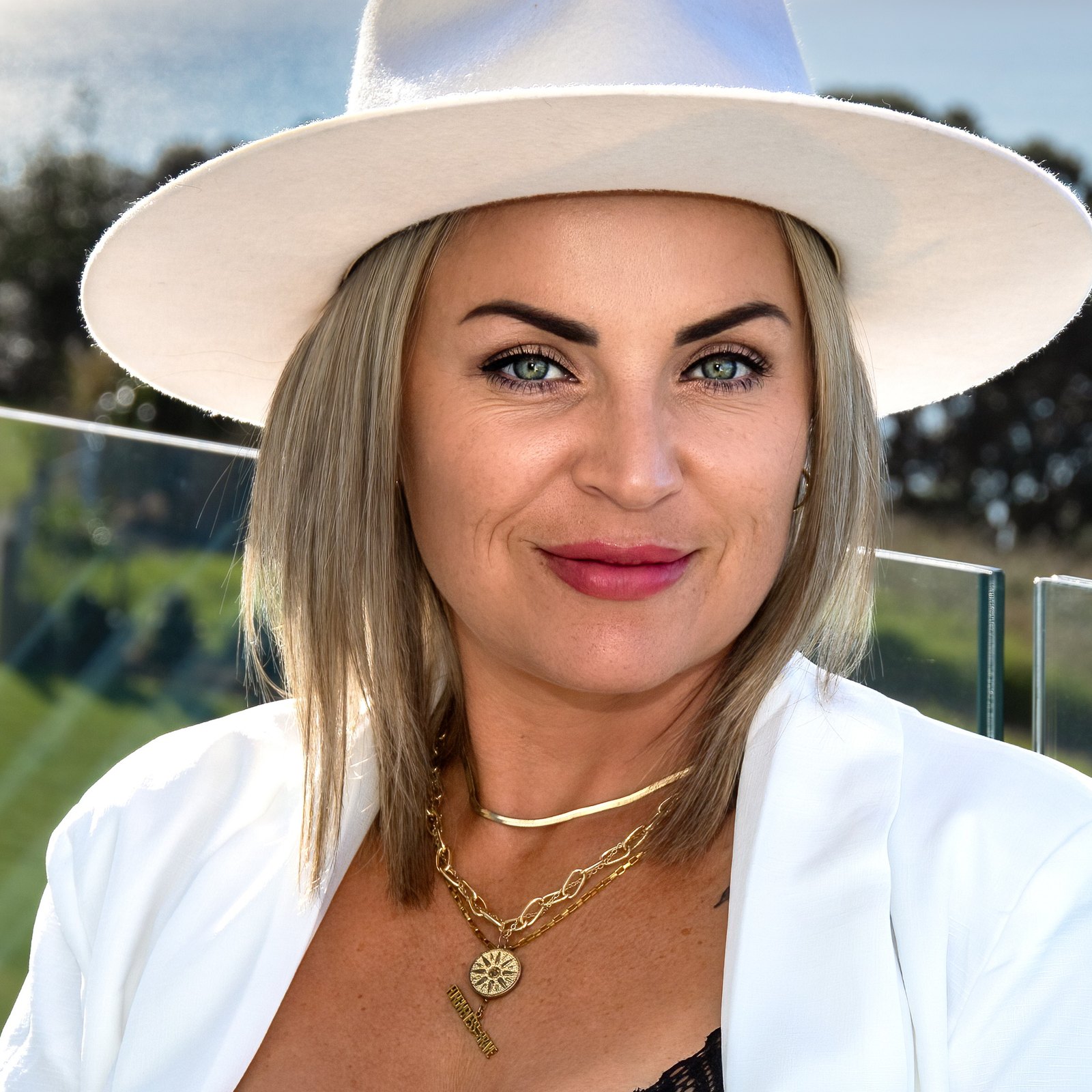 Victoria Bond in a white hat and white blazer, smiling to camera on a terrace above the sea