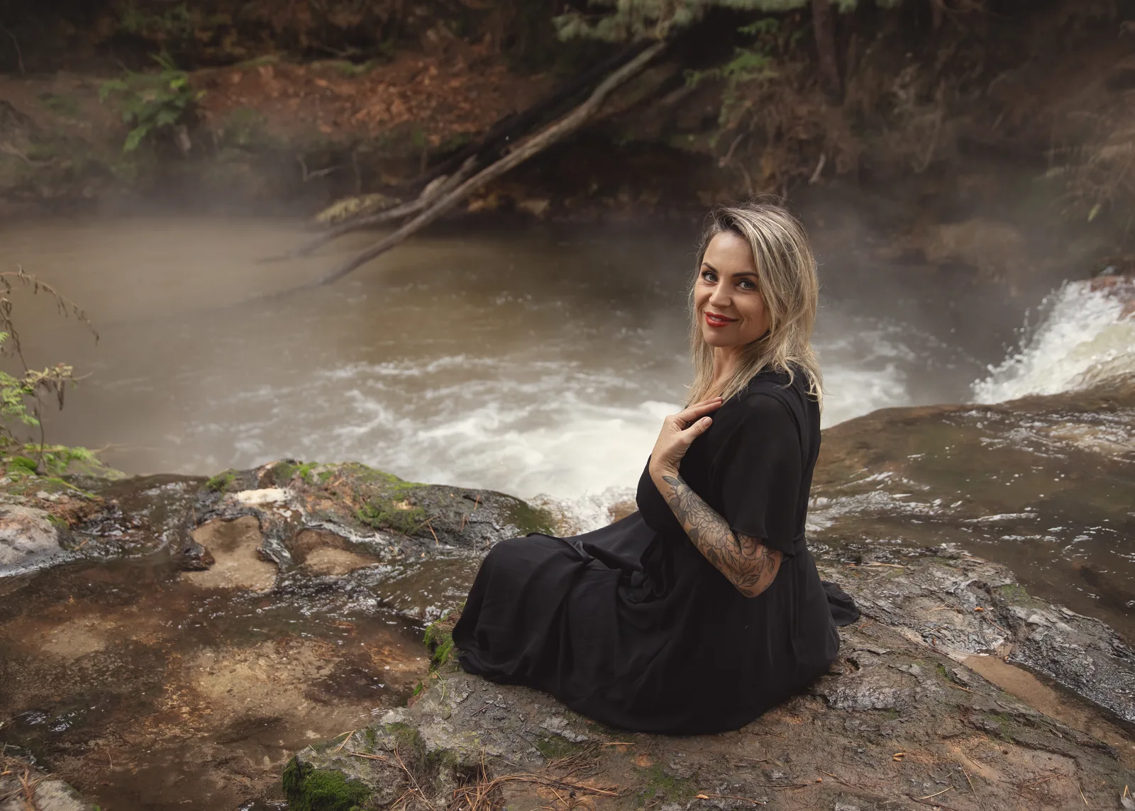 Victoria Bond seated on a rock beside a misty river, looking back at the camera