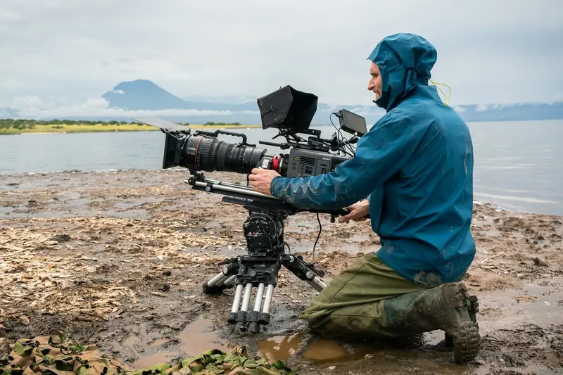 A cameraman kneeling in mud by a lake with a long lens on a tripod