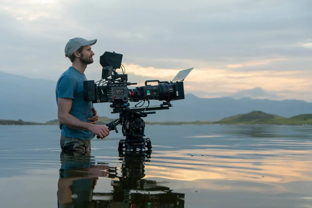 A cameraman standing in a lake at sunset with a cinema camera on a tripod