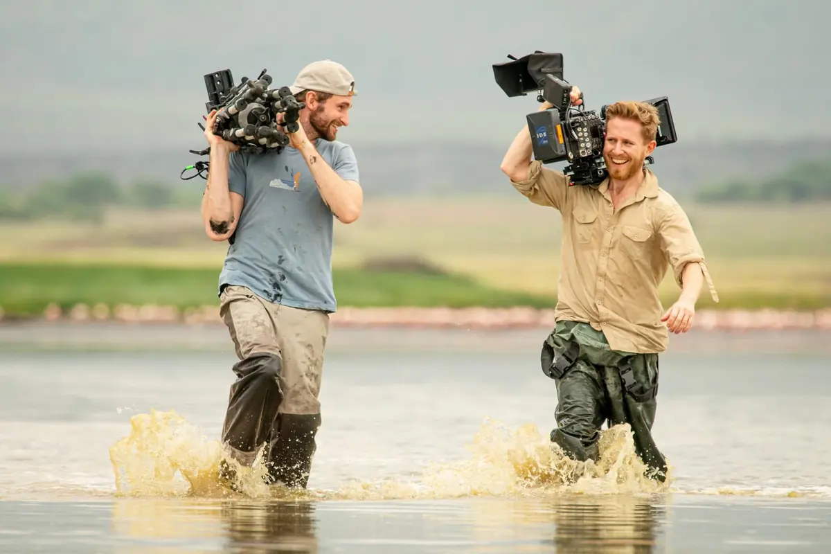 Sebastian and Duncan wading through a shallow lake, each carrying a camera on his shoulder and laughing