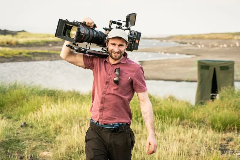 A cameraman walking through grass with a camera on his shoulder