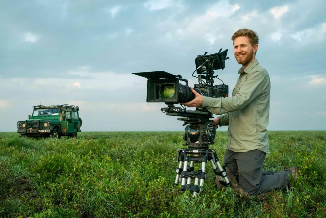 Sebastian kneeling beside a cinema camera on a tripod on a grass plain