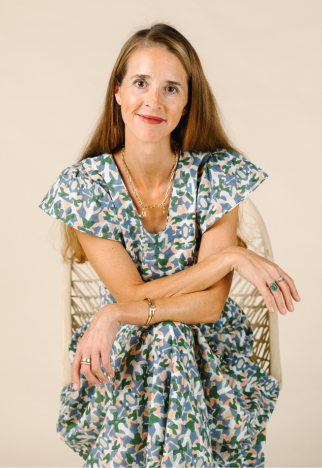 A woman seated on a chair with arms gently crossed, wearing a patterned dress against a neutral background.