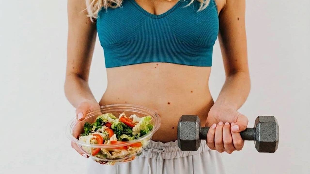 A woman wearing a teal sports bra and white pants holds a bowl of fresh salad in one hand and a dumbbell in the other, set against a plain light background.