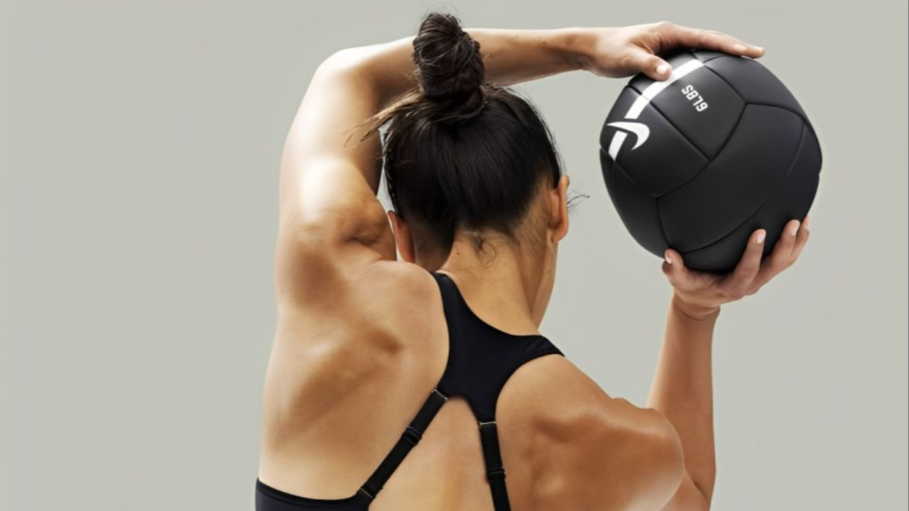 Woman in black workout clothing holding a weighted ball overhead, showcasing strong back and shoulder muscles during a functional strength workout focused on building muscle, bone density, and longevity in a women-only gym environment.