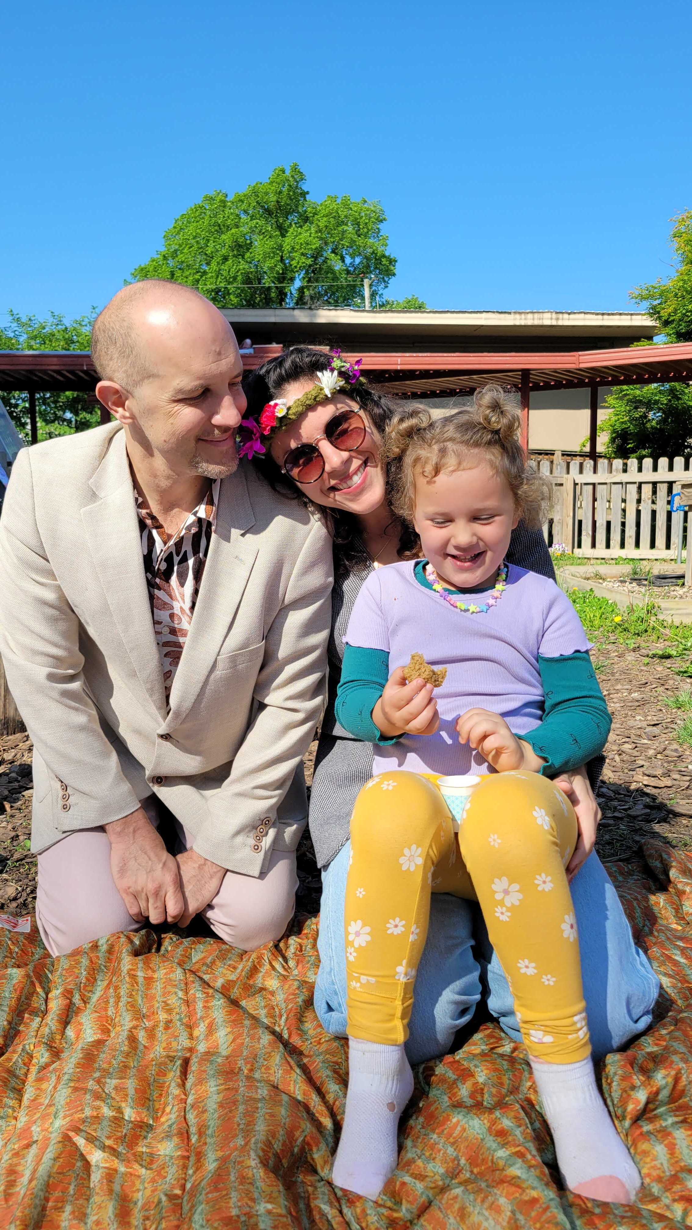Courtney in a flower crown, laughing with her husband and daughter on a picnic blanket