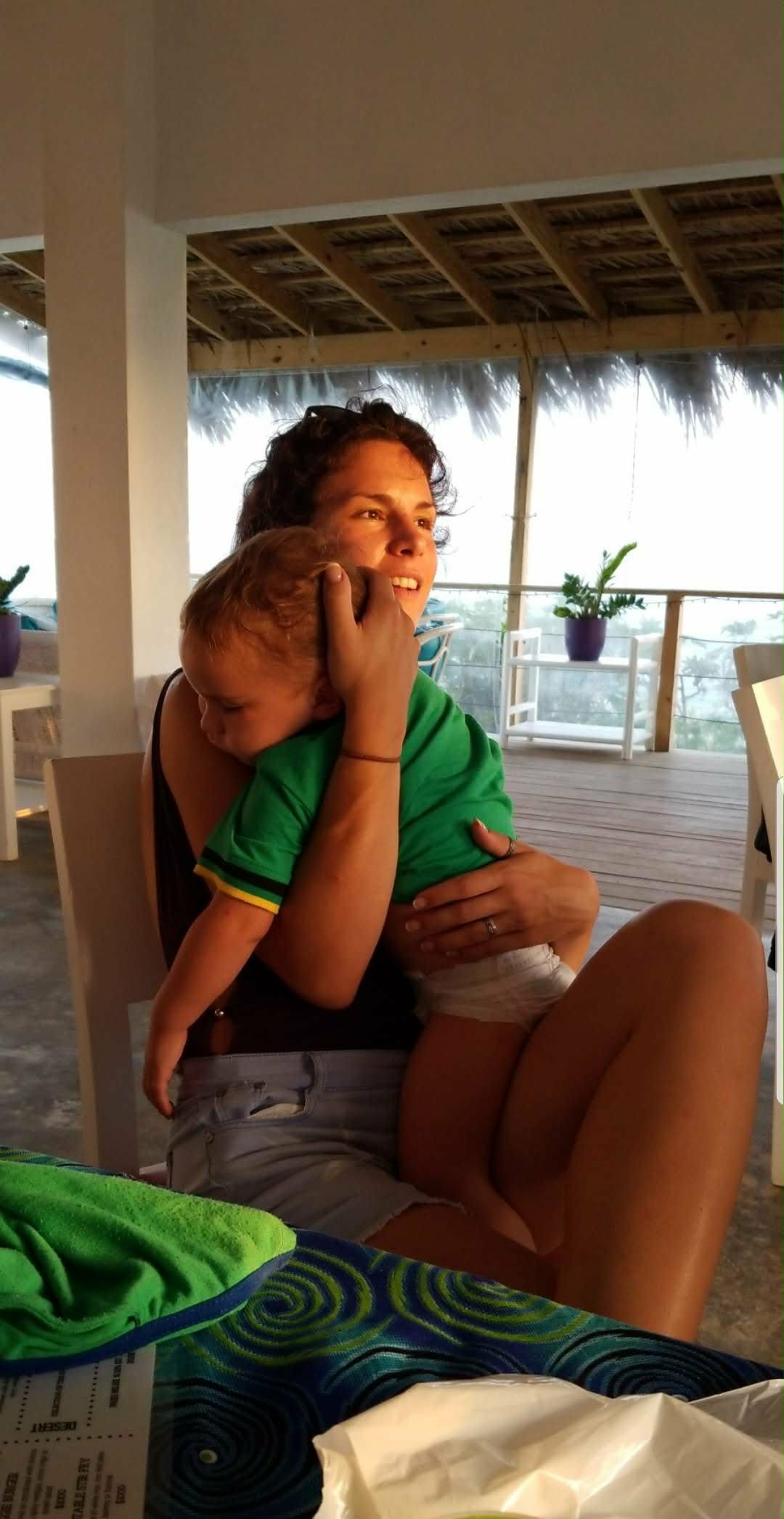 Courtney smiling as she holds her sleeping baby on an open-air porch by the sea