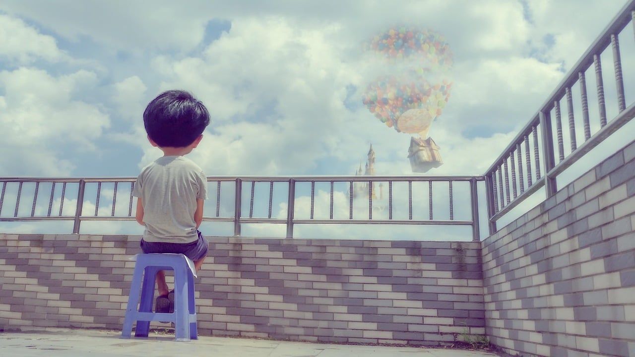 A young boy sits on a stool on a rooftop, gazing at a dreamlike floating house carried by colorful balloons in the cloudy sky.