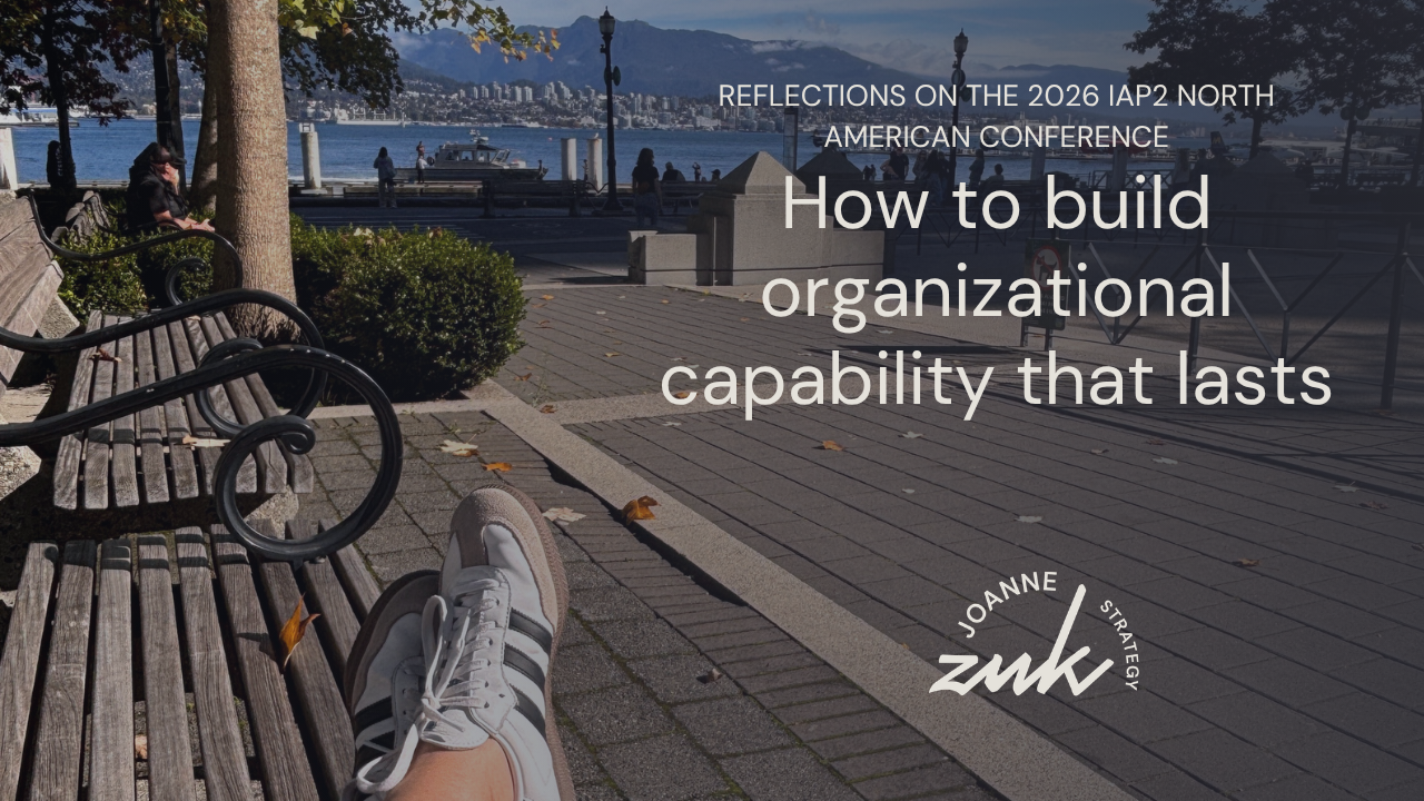 A photograph is taken from the perspective of Joanne, who is sitting on a bench with her feet extended in front of her. The view is of the seawall of Vancouver, looking across to the North Shore. The photo was taken during a break in the IAP2 North American Conference. 