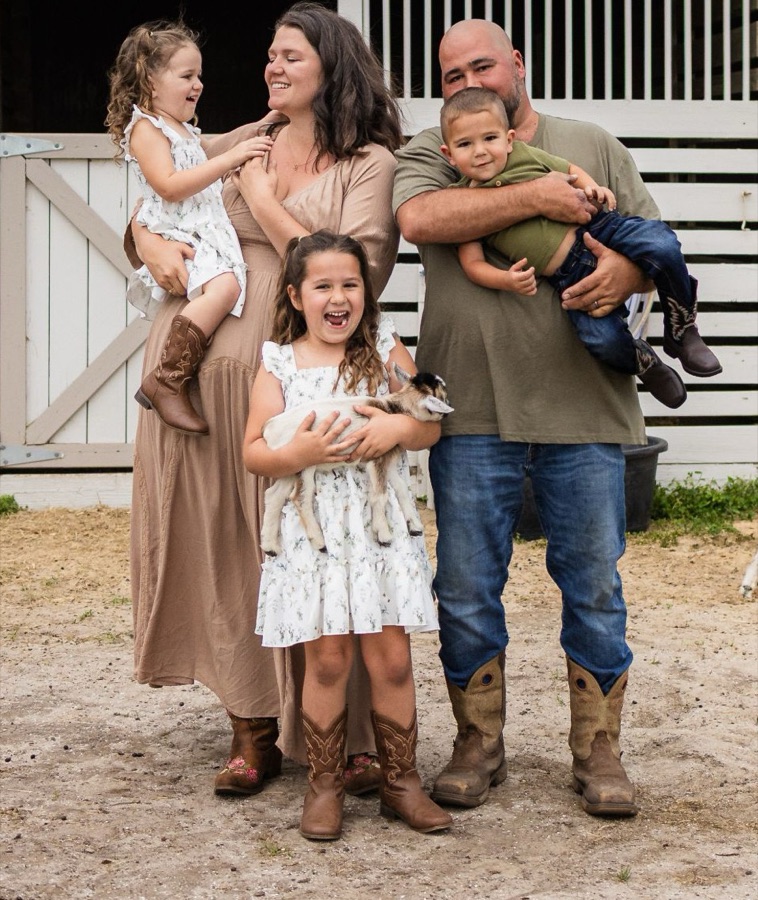 Taylor, her husband and their three kids laughing in front of the barn, holding a baby goat