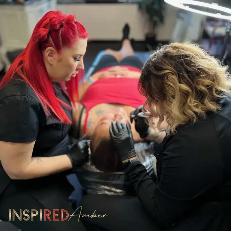 PMU educator Amber providing hands-on instruction and supervision to a student practicing eyebrow tattooing on a live model.