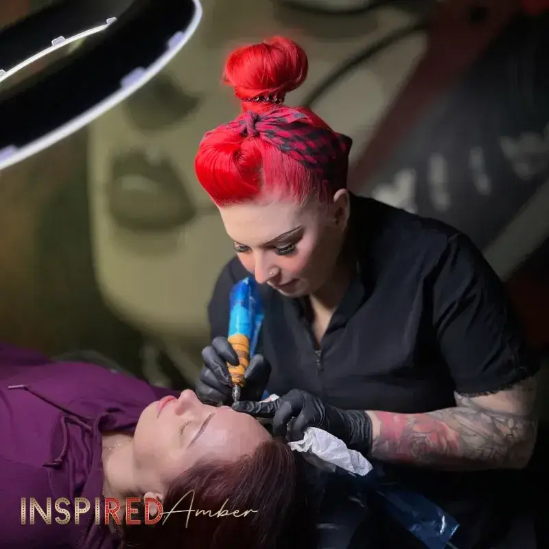 Permanent makeup instructor Amber with red hair using a PMU machine tattoo pen to perform a live eyebrow tattoo demonstration on a client.