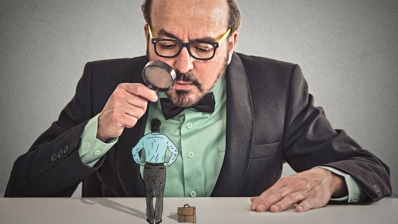 Man with a magnifying glass looking down at a miniature businessman and a tiny briefcase