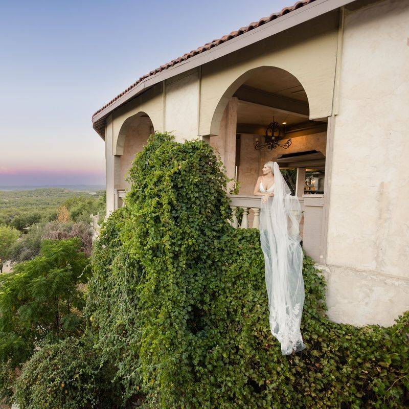 Bride in a flowing veil looking out from a balcony at sunset