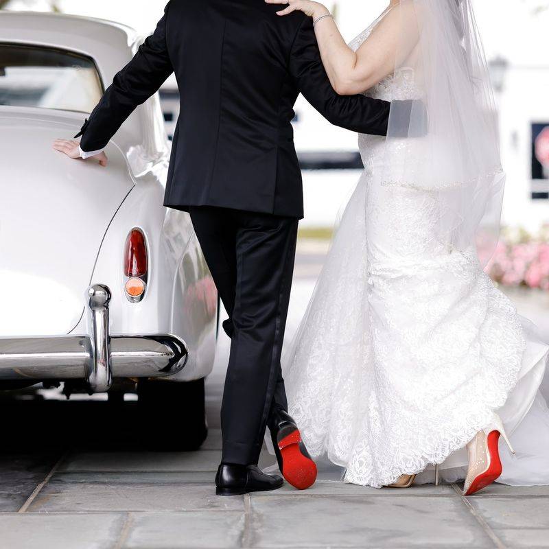 Bride and groom walking hand in hand beside a vintage white getaway car with a 'True Love' license plate