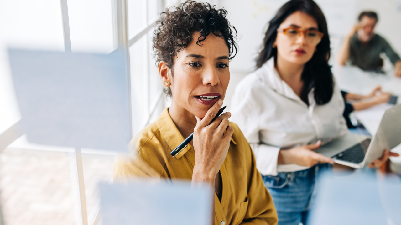 A focused woman in a yellow shirt thinking thoughtfully during a meeting with colleagues, representing the Inner Empire™ truth that high-performing women need systems not willpower to protect the energy that makes their leadership possible.