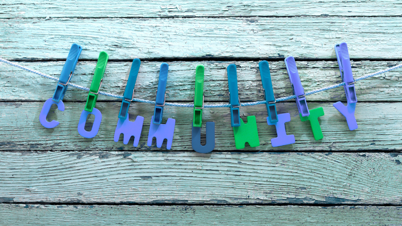 The word "community" spelled out in colorful foam letters clipped to a clothesline against a weathered teal wood background.