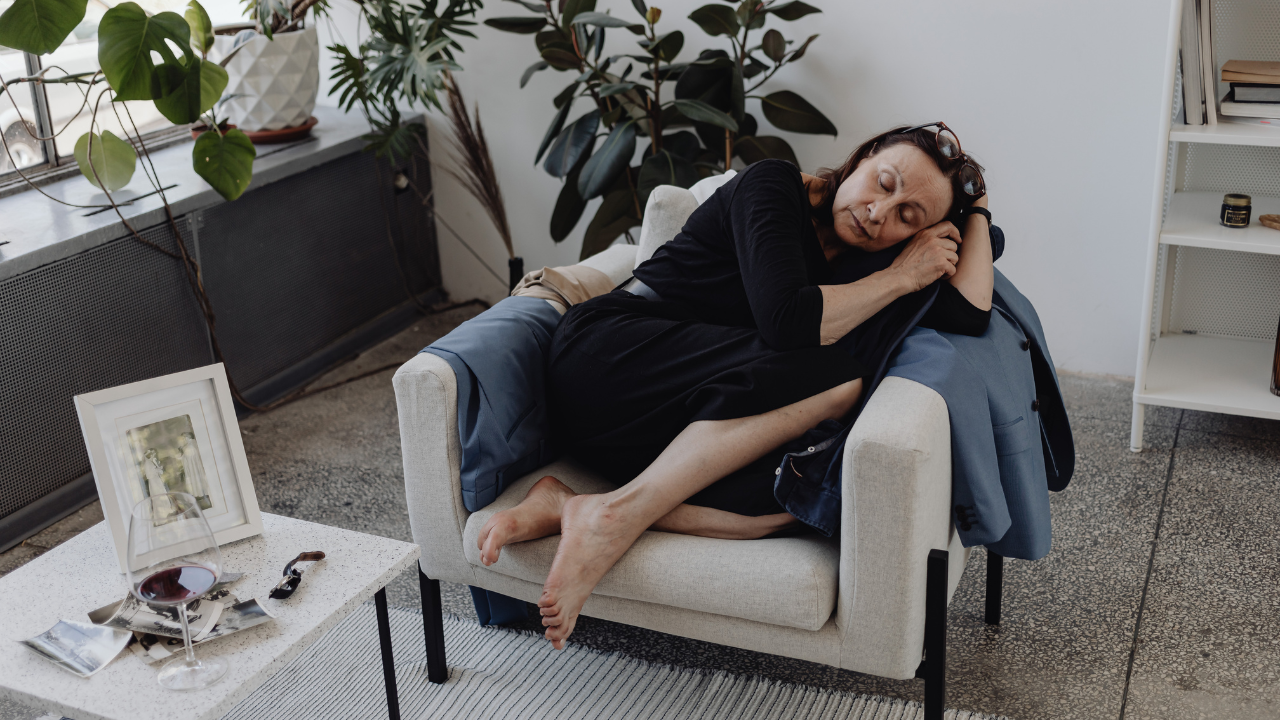 A woman in work clothes asleep in an armchair, one shoe off, with a wine glass and old photographs on the table beside her, representing physical exhaustion from misalignment.