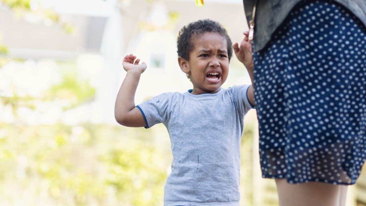 Toddler outdoors with one arm raised, standing next to a parent