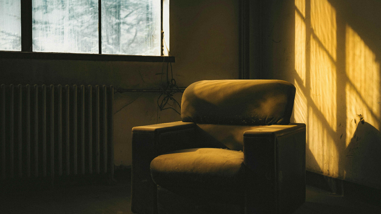 A worn armchair sits empty in warm golden light by a window, casting long shadows across a bare room.