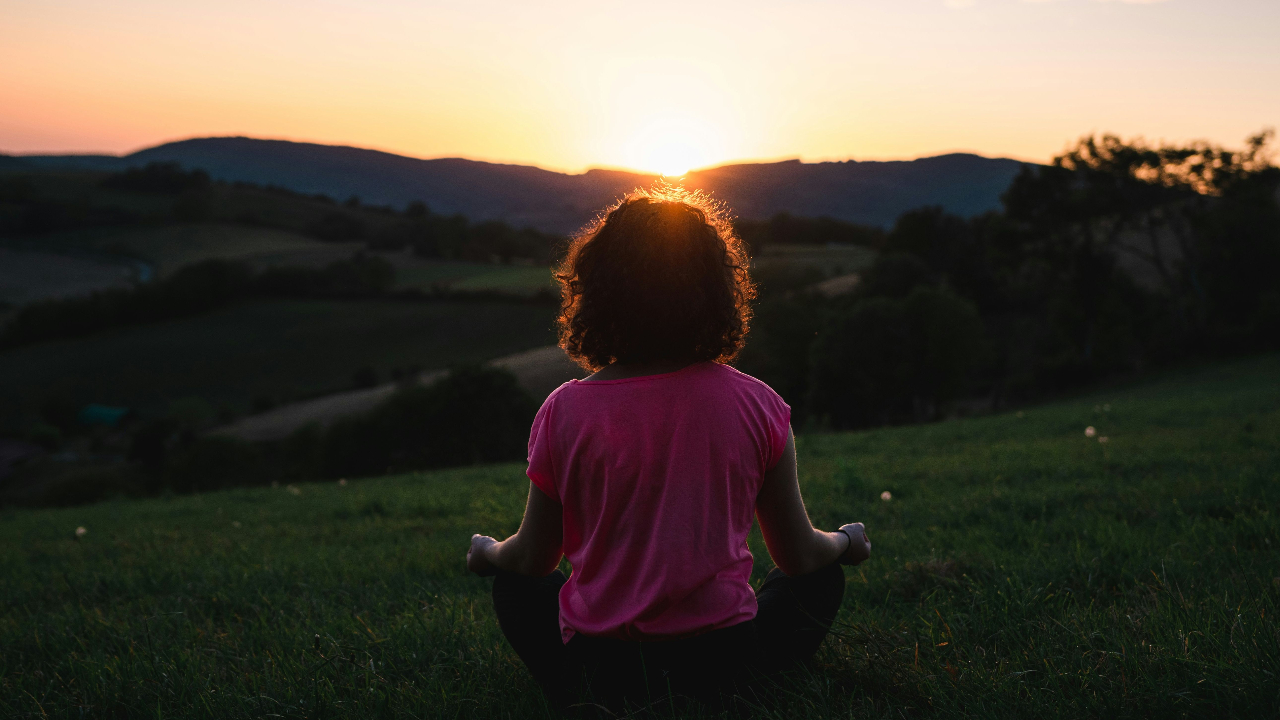 A person sits in a meditation pose on a grass hillside, facing away from the camera toward a warm sunset over rolling mountains.
