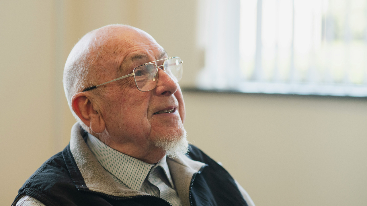 "An older man with glasses and a white beard, smiling and looking up while listening, seated near a sunlit window."