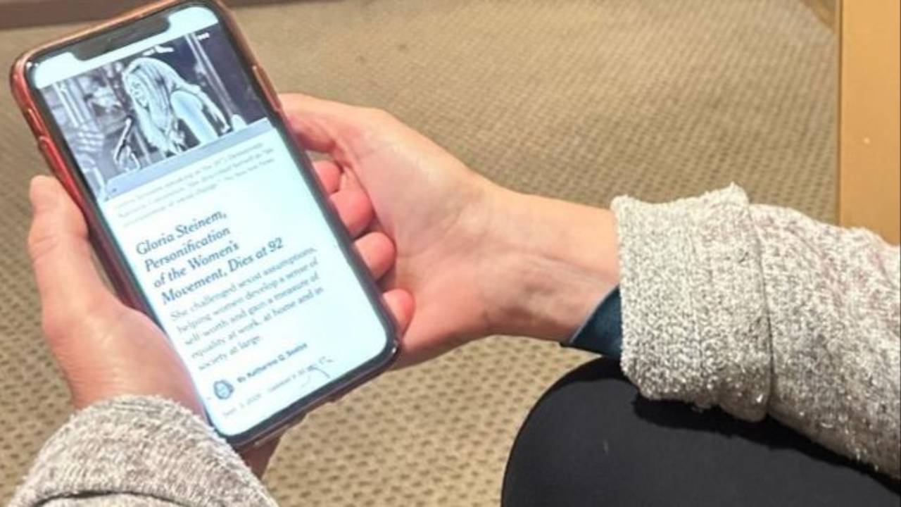 A person sitting cross-legged reads the Gloria Steinem obituary on their phone, the headline "Gloria Steinem, Personification of the Women's Movement, Dies at 92" visible on screen, hands cradling the phone.
