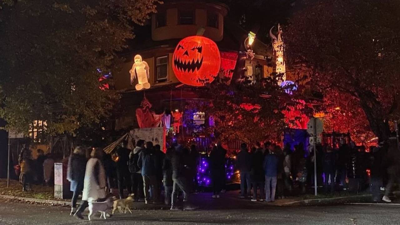 A skeleton draped in cobwebs sits on a wooden garden bench while trick-or-treaters visit a candlelit, decorated porch behind it.