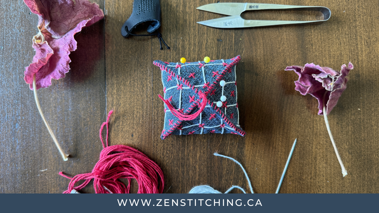 A number of sewing tools are sitting on a wooden tabletop, with a couple of dried pink flowers. At the centre is a pin cushion with a number of pins in it; surrounding it are a pair of sharp steel snips, a palm thimble, a threaded Sashiko needle, and some red Sashiko thread. A blue bar across the bottom reads "www.zenstitching.ca"