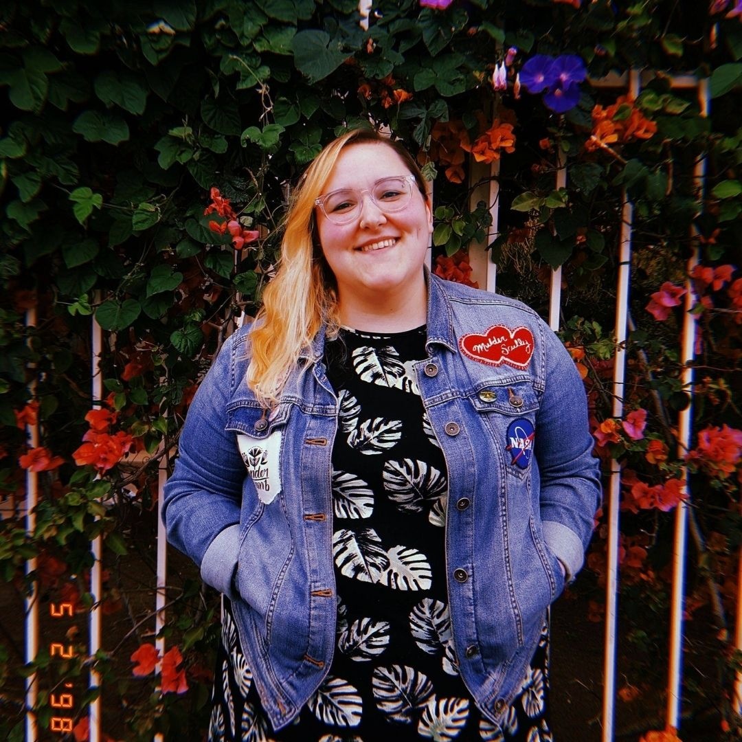 Photo of Sabrina Volante, Graphics Designer. Volante stands in front of a fence that has flowers on it while wearing a floral dress and a jean jacket.