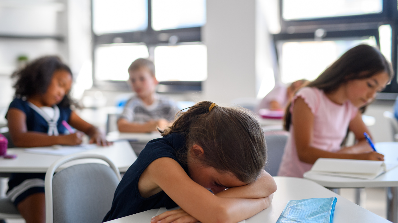 Young student resting their head on a desk in a classroom while other children continue working at their desks.