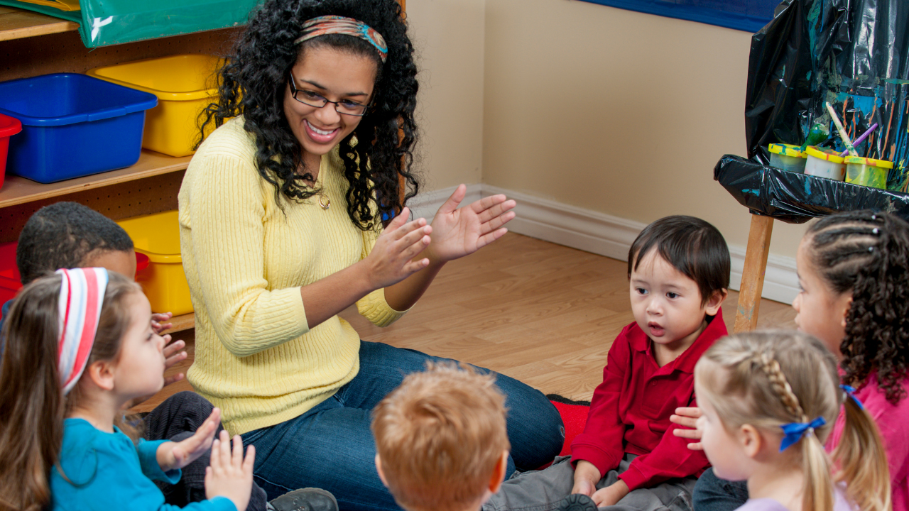 Preschool teacher sitting on the floor clapping hands during a circle time activity with a diverse group of young children.
