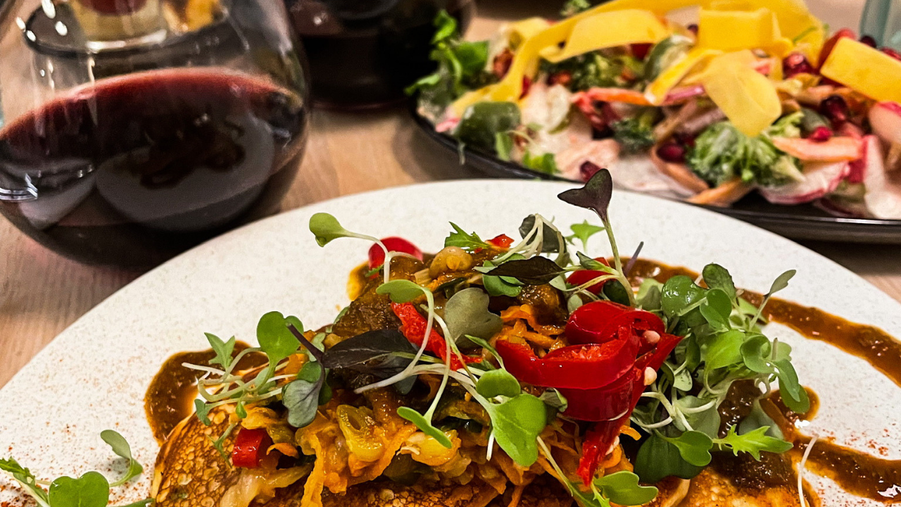 A plated savoury dish with microgreens and roasted peppers, a glass of red wine, and a mixed salad