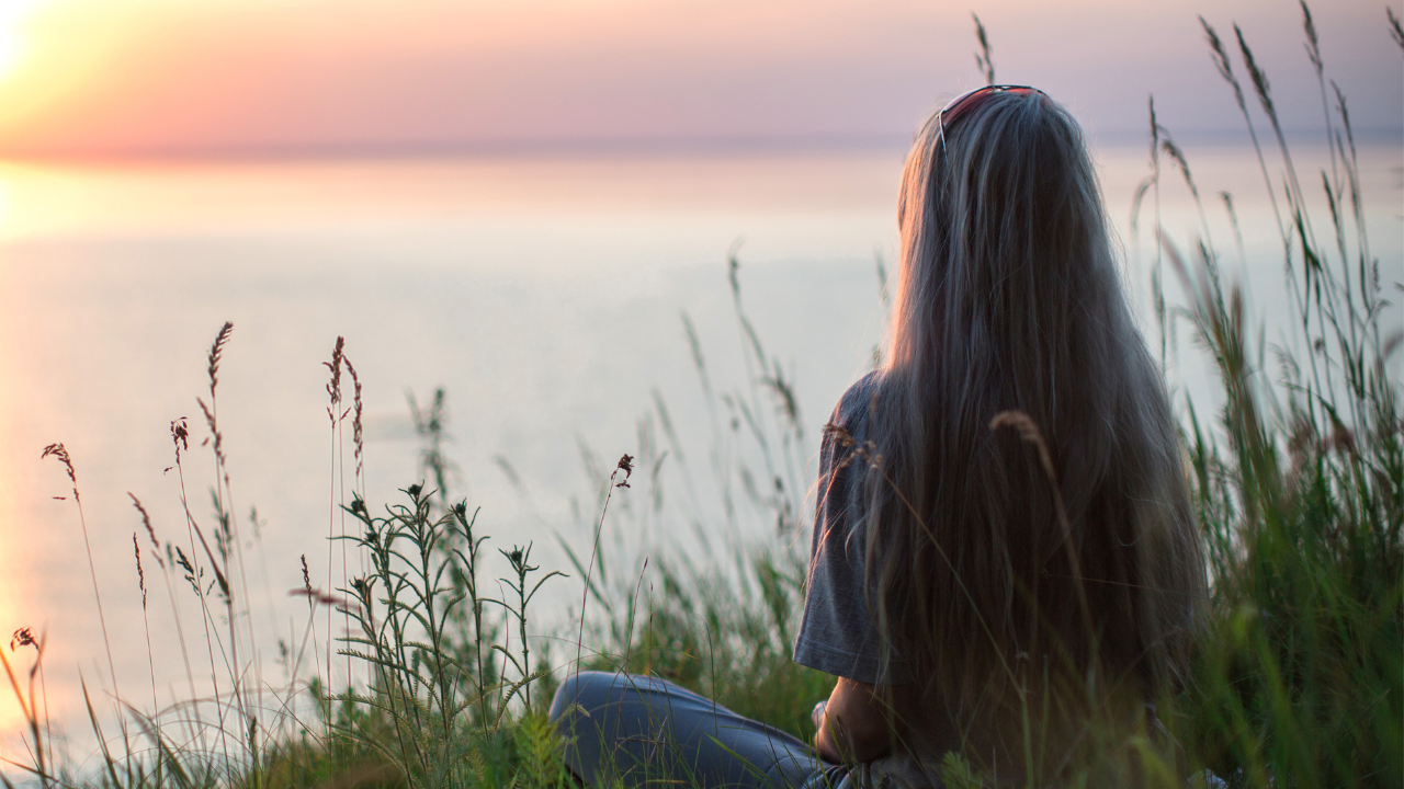 Woman sitting among coastal grasses overlooking calm water at sunset.