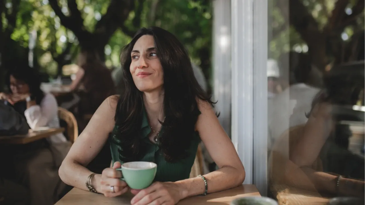 Lindsay Kassem, executive health and mindset coach, sitting at a cafe table holding a coffee cup, looking outward in a reflective moment