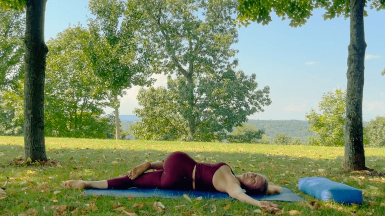 Caitlin lying on a yoga mat outdoors, resting under trees with a peaceful nature view