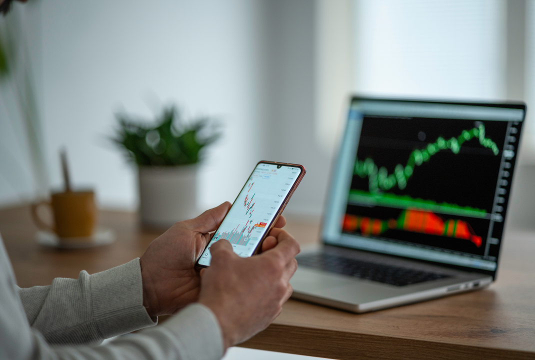 Man using a trading app on a smartphone and laptop displaying a stock market chart with rising and falling candlesticks