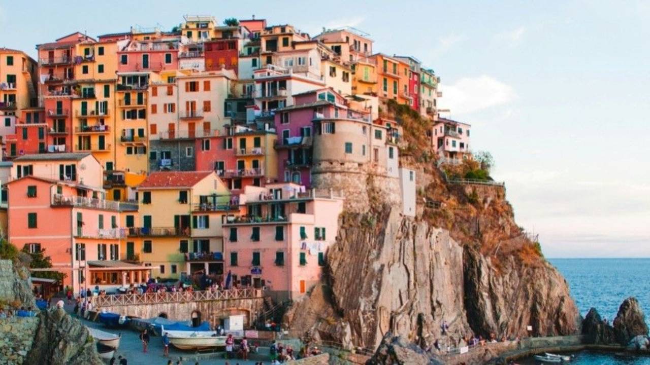 Colourful pastel buildings perched on a steep rocky cliff above the Mediterranean Sea in Manarola, Cinque Terre, Italy, with boats and people gathered along the waterfront.