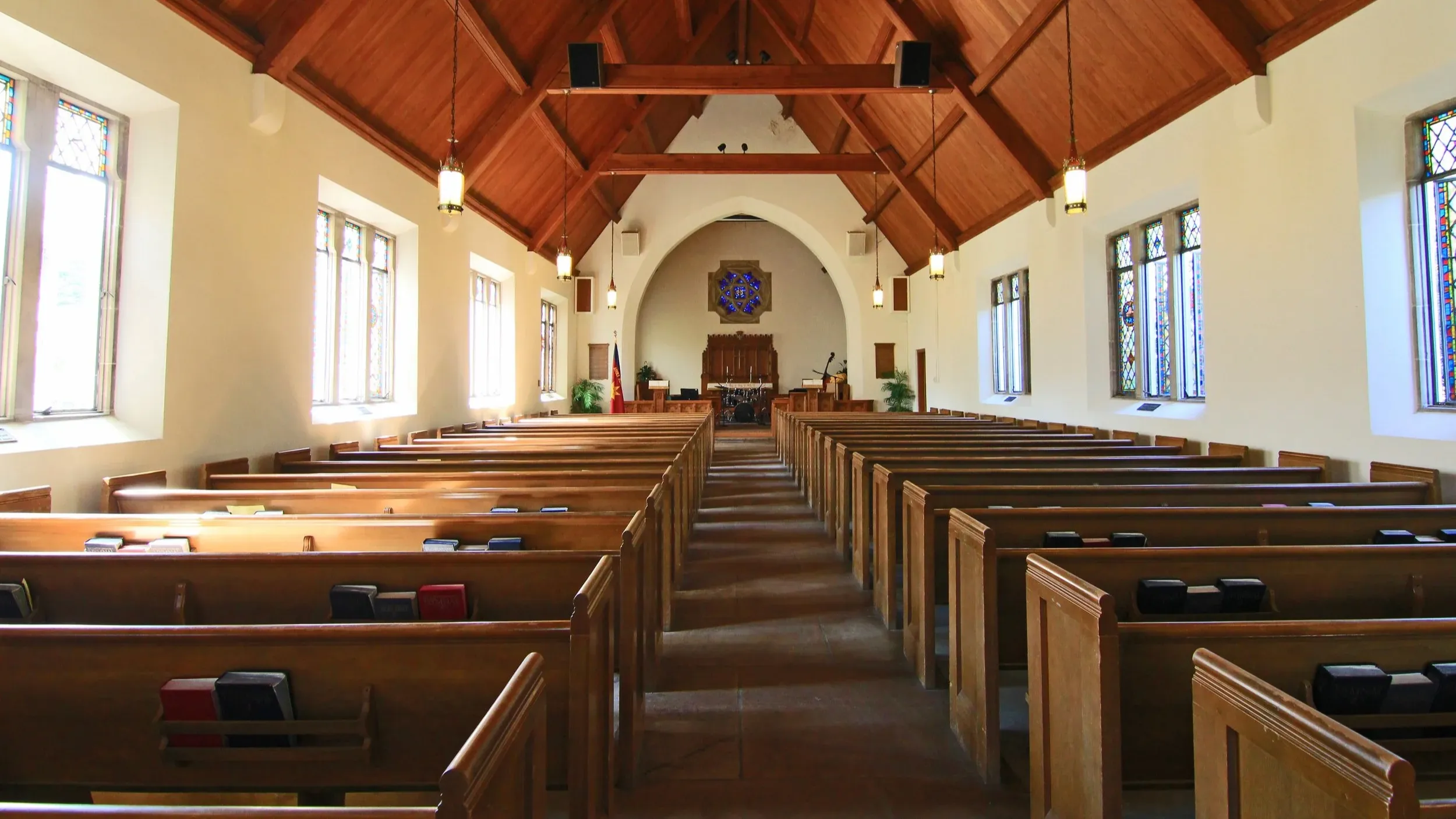 Empty wooden church sanctuary with exposed beam ceiling, photographed from the back showing rows of pews and natural light filtering through high windows.