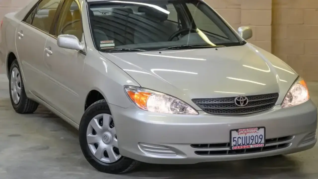 An older silver sedan on a lift in a garage, hood open, while a mechanic looks up from the engine.