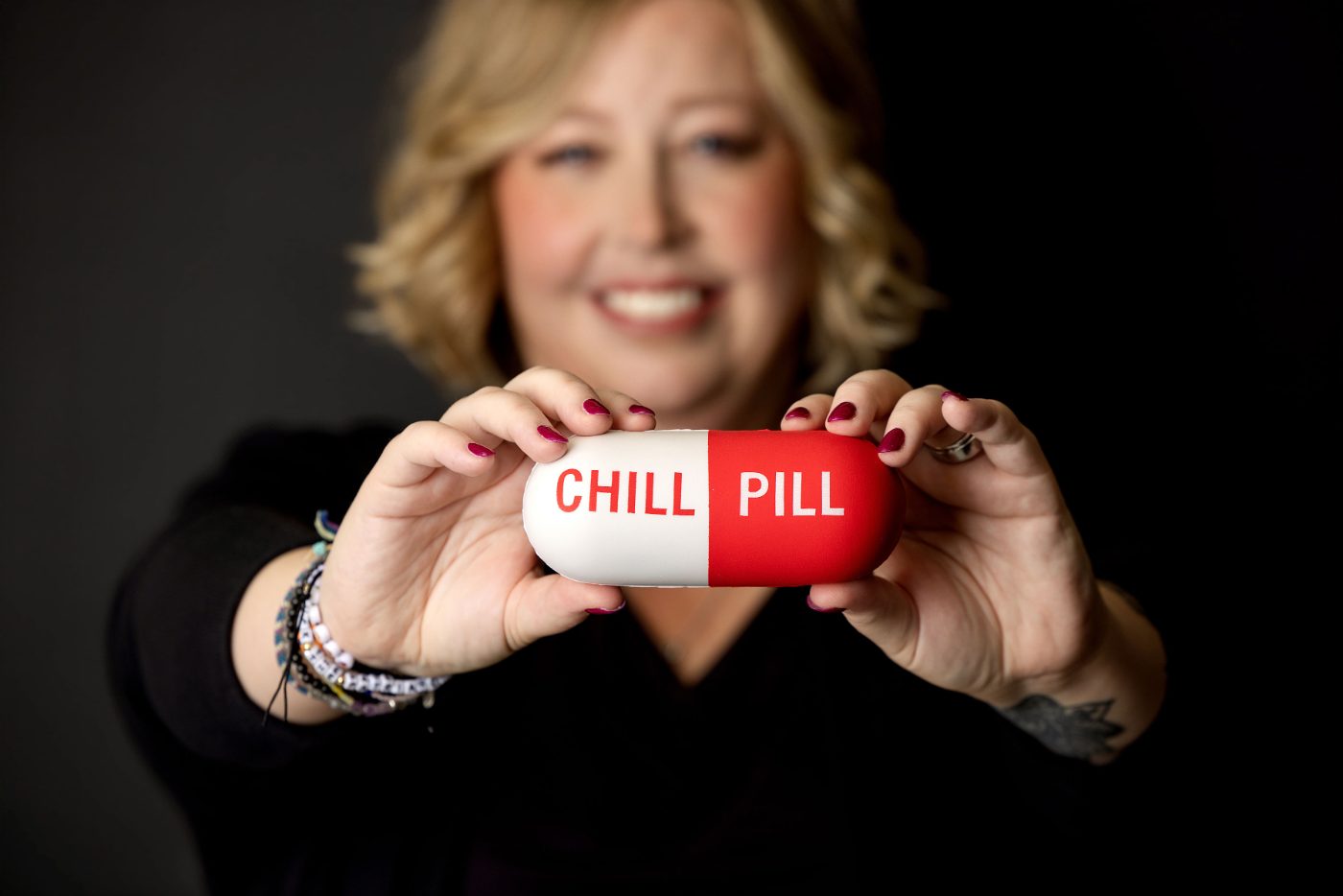 Becca Gerlach smiling and holding a red and white pill-shaped pillow that reads Chill Pill against a black background