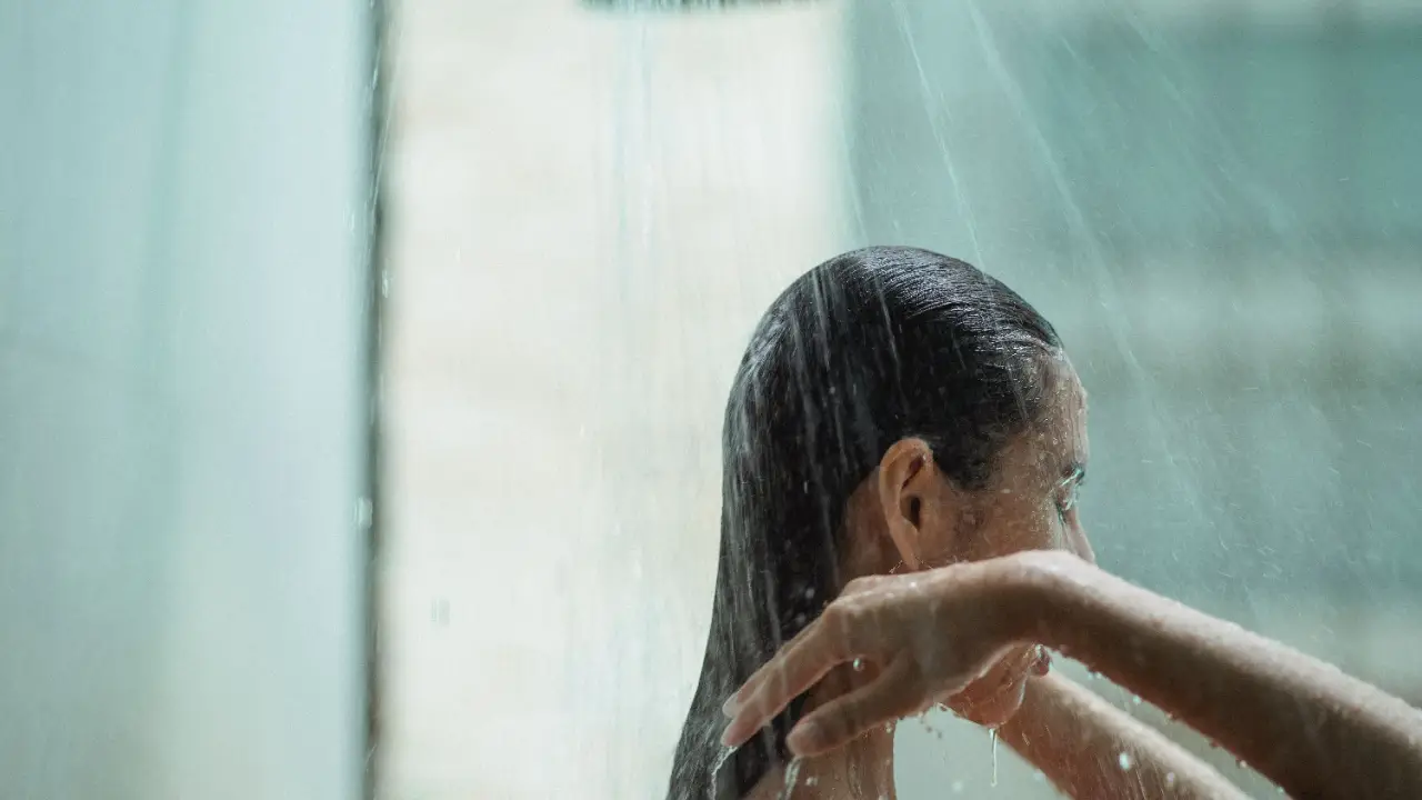 A woman stands under a shower with her back to the camera, water falling around her in soft light.